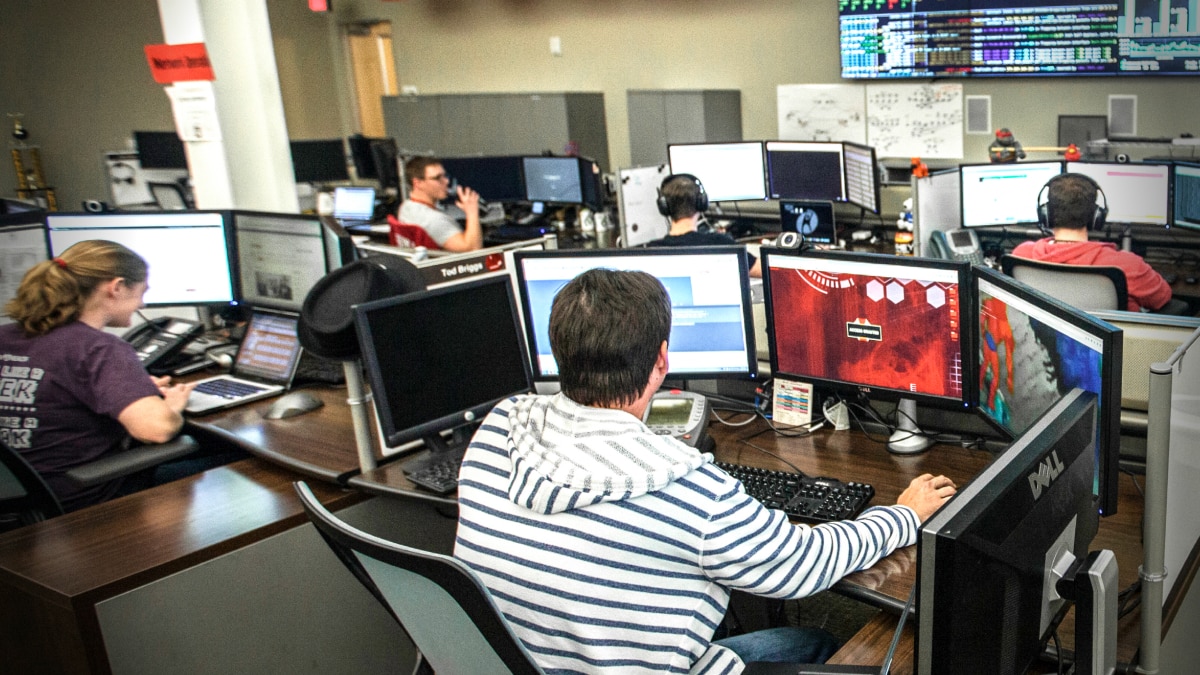Four coworkers gathered at a desk reviewing content on a laptop in a modern office setting.