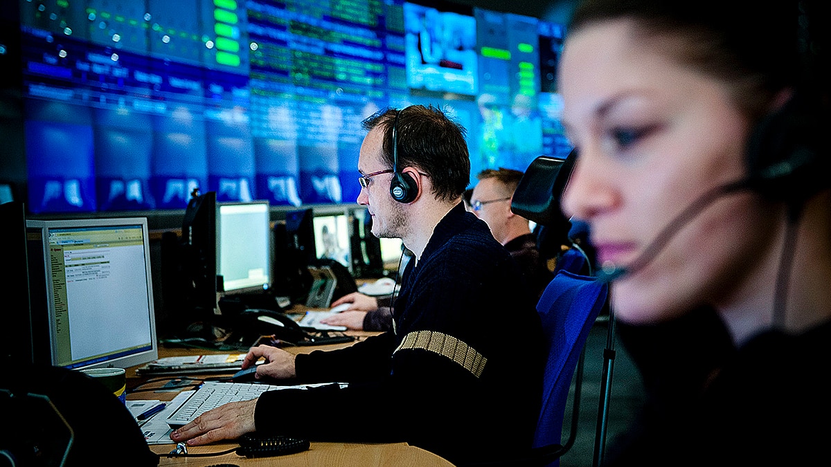 Call center operators wearing headsets at computers, large wall displays showing system data in blue-lit control room