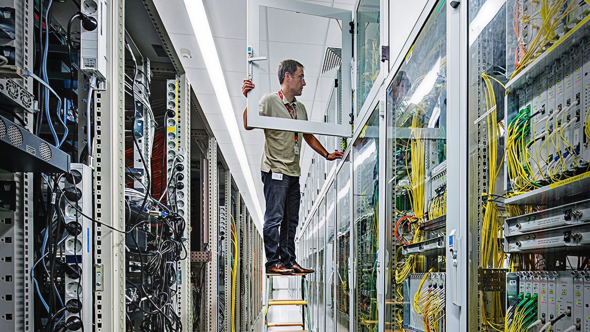 Technician standing on ladder servicing network server racks with visible cables and equipment in data center aisle