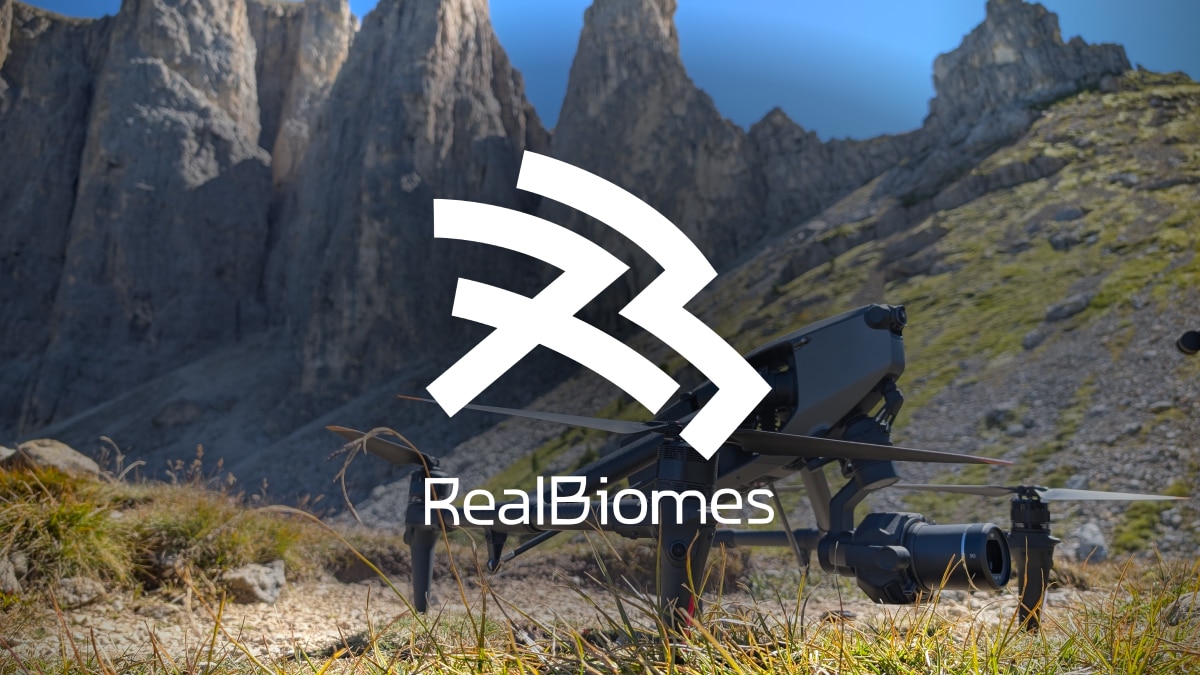 Drone with camera on grassy alpine ground before tall rocky peaks; white RealBiomes logo and text “RealBiomes” overlay.