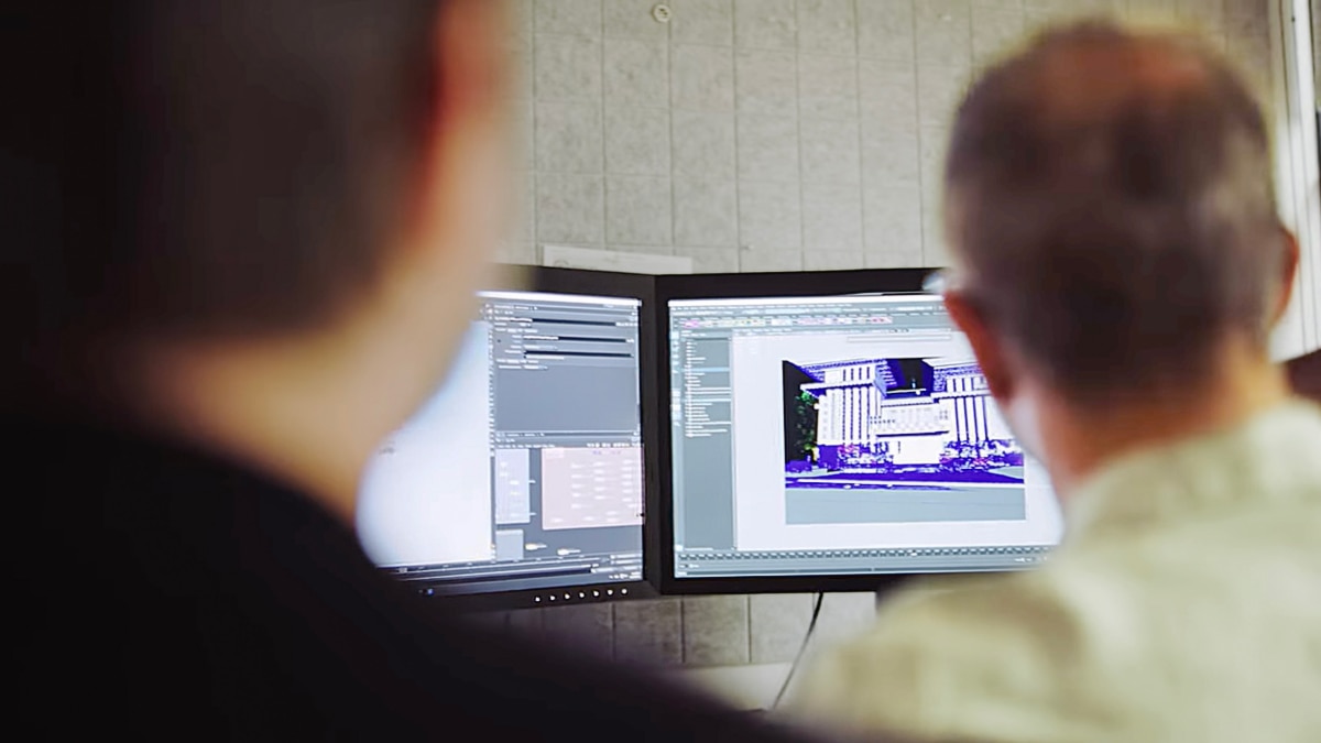 Two people view dual monitors showing 3D modeling software with a building render and interface panels in a studio setting