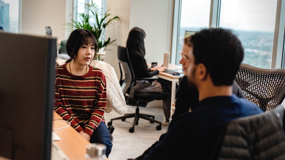 Three coworkers talking in a bright office while another works at a desk in the background
