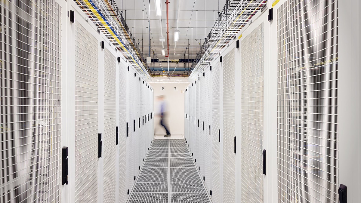 Long aisle of white server racks in a data center with overhead cables and a blurred person walking