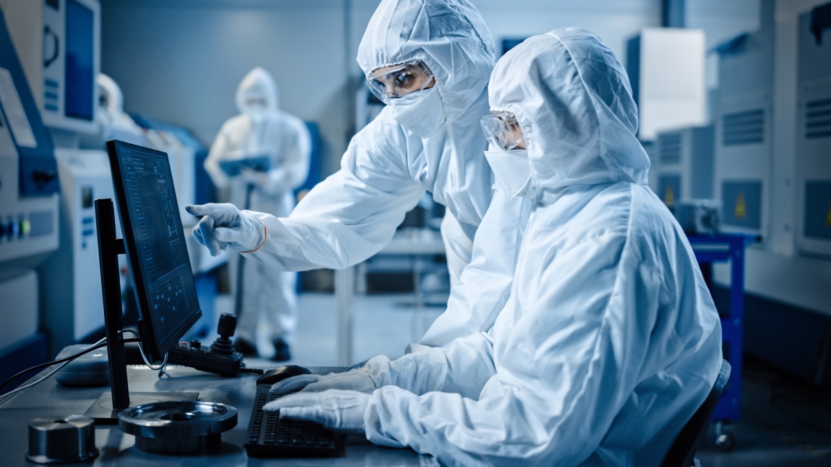Two cleanroom engineers in protective suits analyze data on computer screen in semiconductor lab