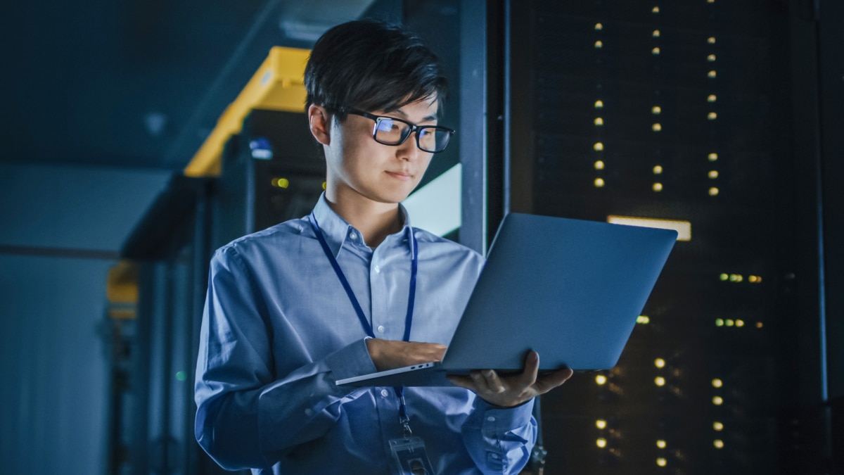 Engineer with glasses uses laptop in server room with illuminated racks in background