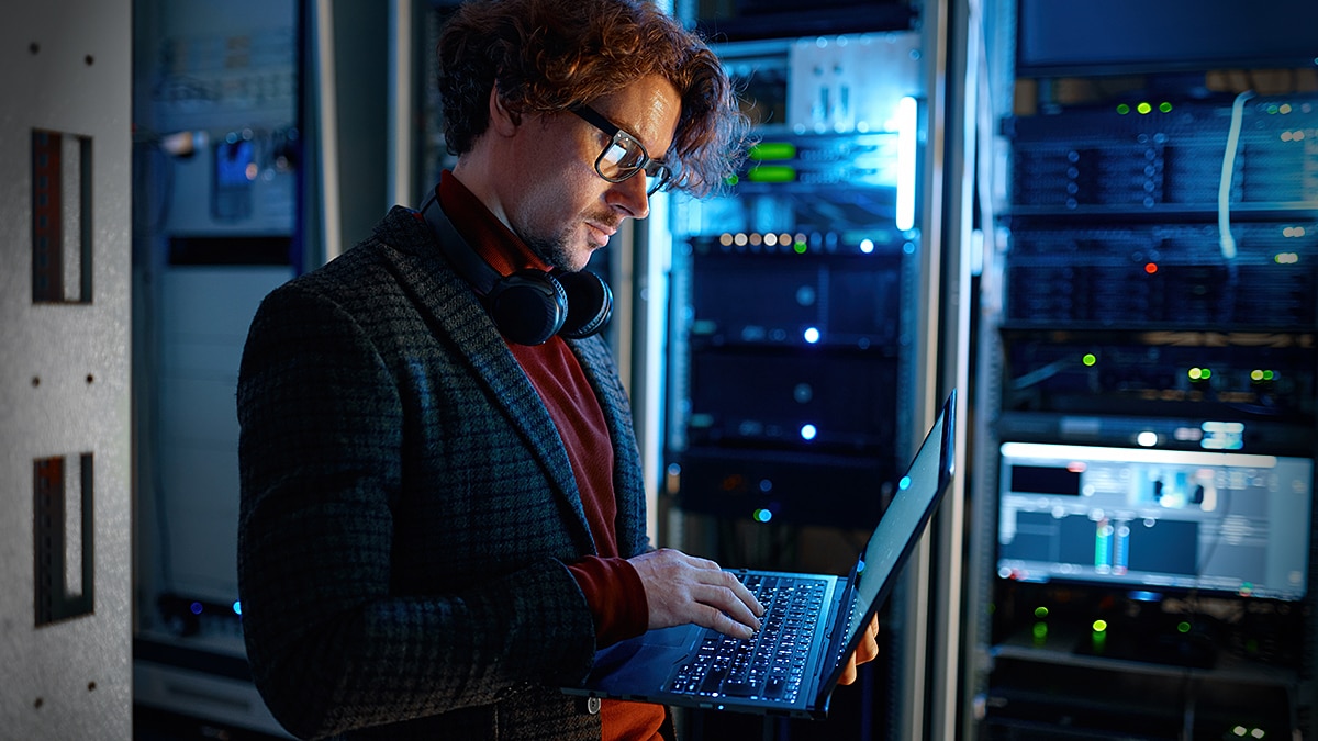 IT professional using a laptop in a server room with illuminated data center racks.