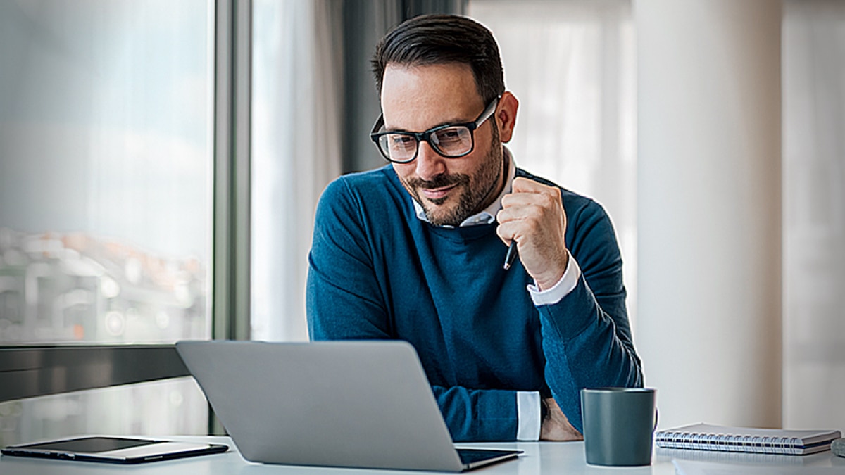 Man wearing glasses working on a laptop at a desk with coffee mug and notebook nearby.