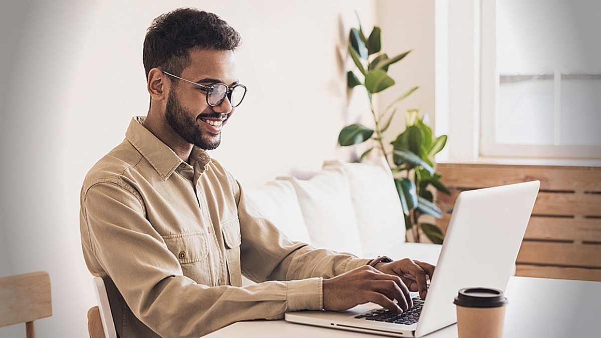 Smiling professional using a laptop at a bright home office desk with coffee cup nearby.