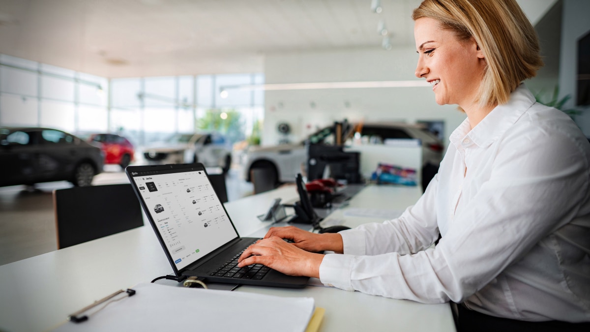 Woman using dealership software on a laptop inside a bright car showroom