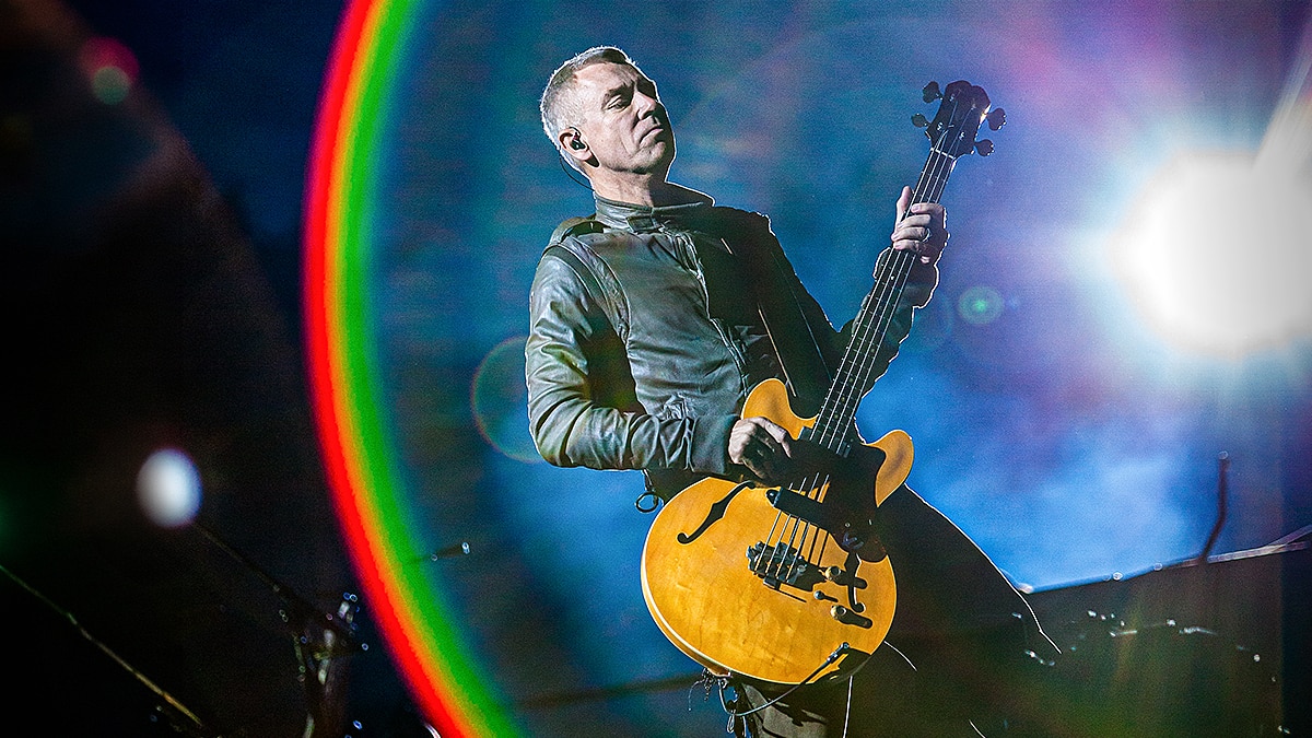 Musician playing a yellow bass guitar on stage with bright lights, smoke, and a rainbow lens flare.