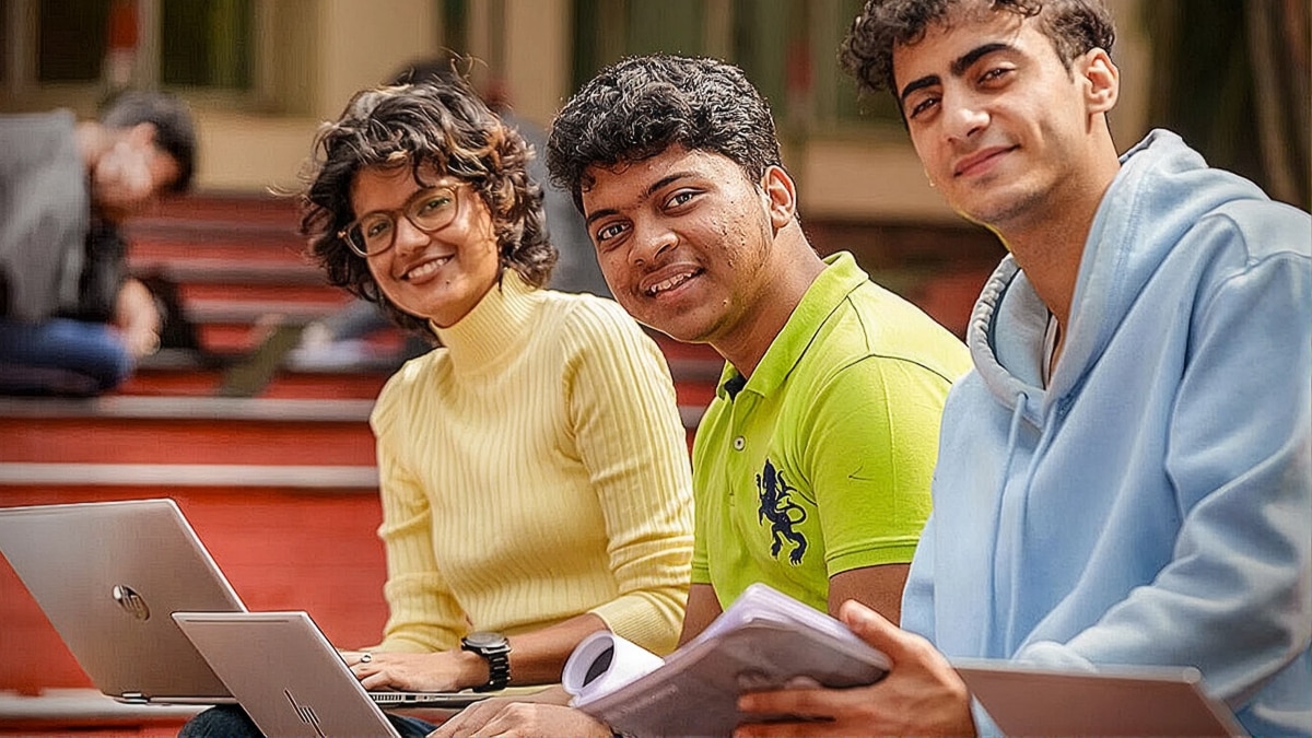 Three students seated outdoors with laptops and notebooks, smiling while studying together on campus.