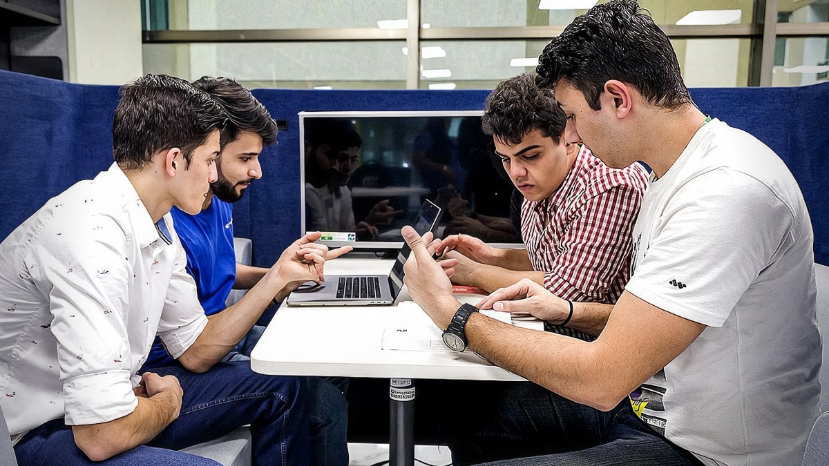 Four students collaborating around a table with laptops and mobile devices in a campus study area.