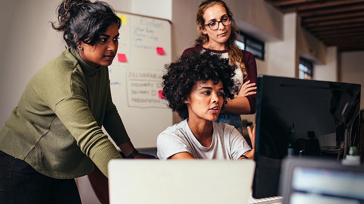 Three developers collaborate around a desktop monitor in an office, with notes and a whiteboard behind them.