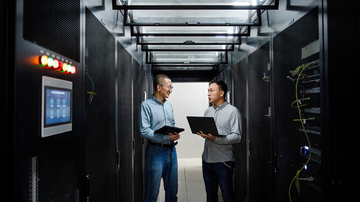Two engineers discuss systems while holding a laptop and tablet between black server racks.