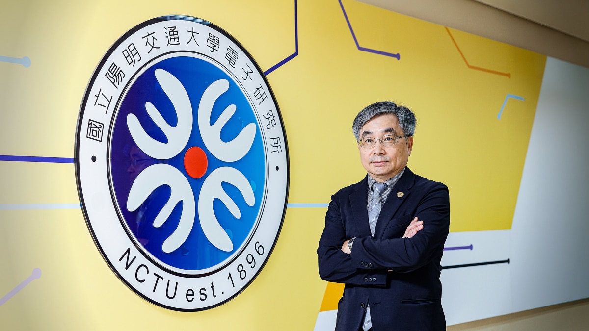 Man in a suit stands beside the blue and white NCTU est. 1896 university emblem on a yellow wall.