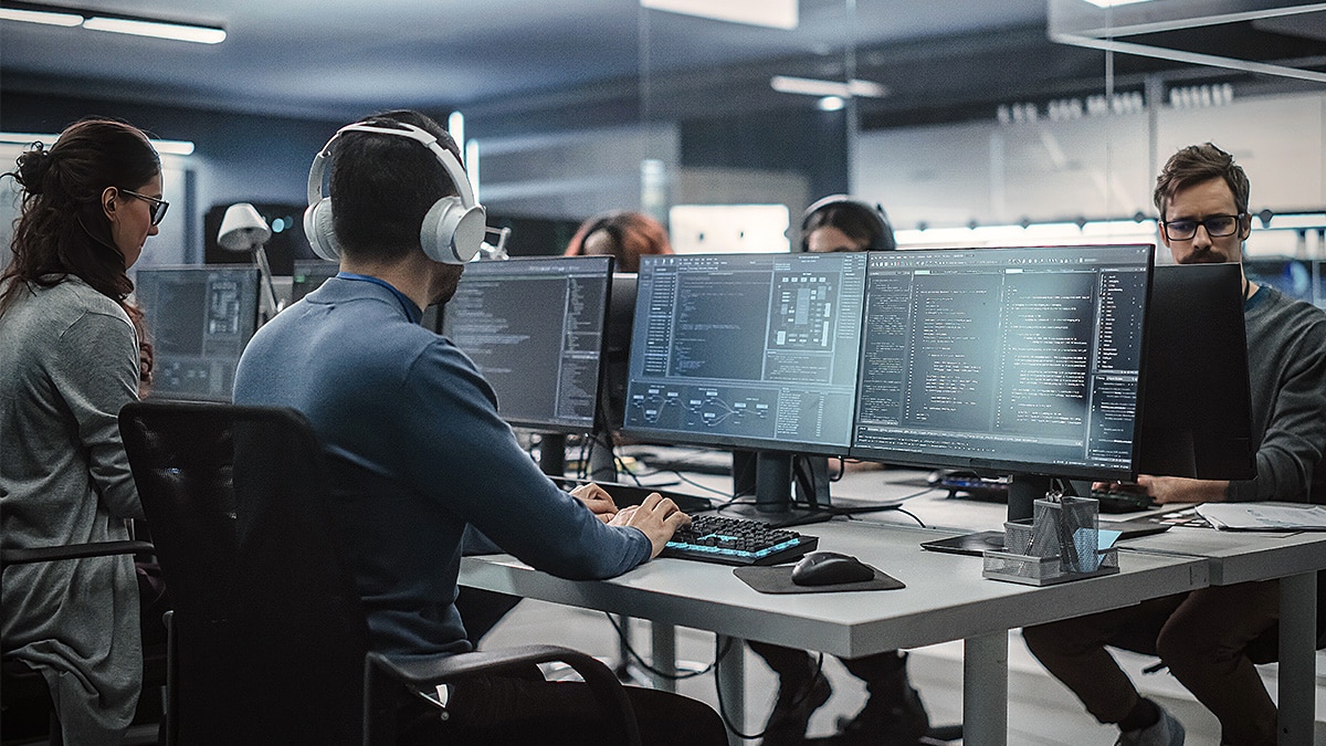 Developers work at multi-monitor stations displaying code and software tools in a modern office.