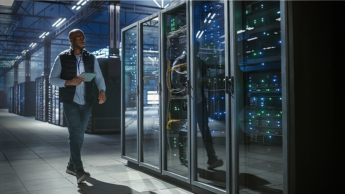 Technician holding a tablet walks beside illuminated server racks in a large data center.