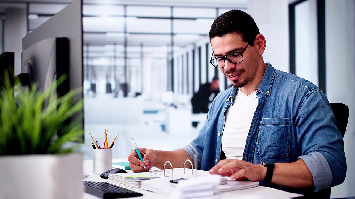 Person wearing glasses and a blue denim shirt writes in a binder at a desk in a bright modern office.