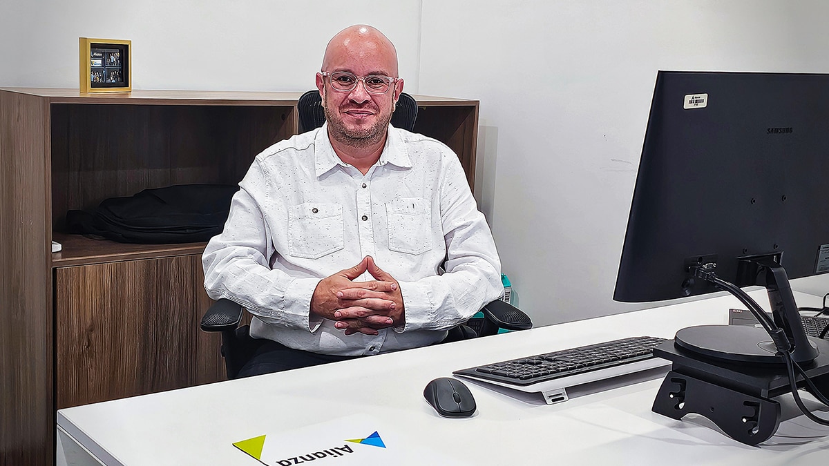Person in a white shirt and glasses sits at an office desk with a Samsung monitor and visible Alianza logo.