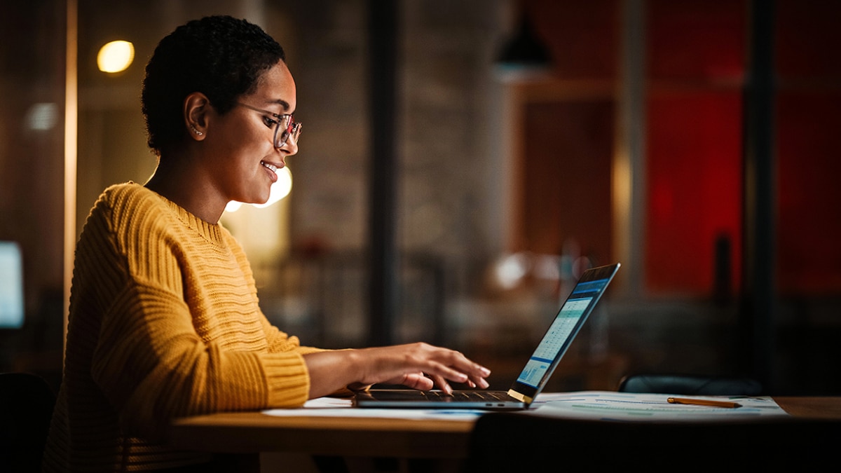 Person wearing glasses and a gold sweater works on a laptop at a desk in a warmly lit office.