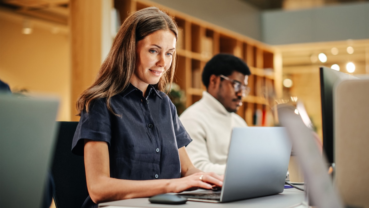 Two professionals work on laptops at desks in a modern office.