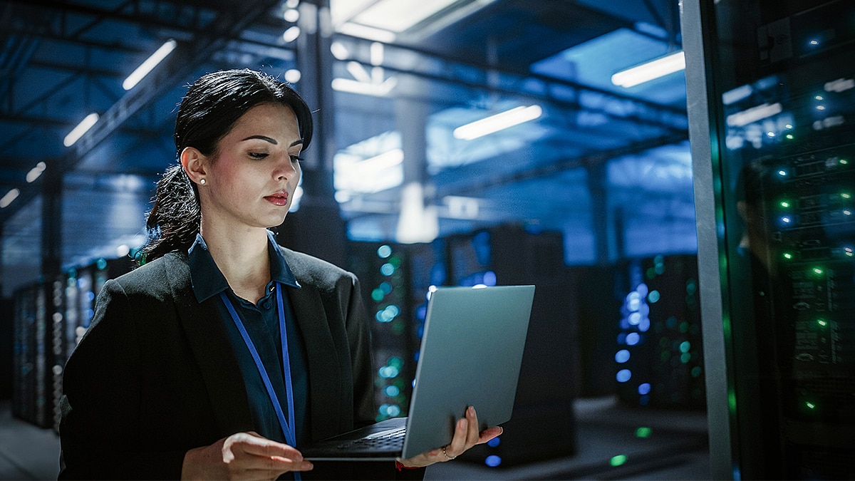 Woman in a black blazer using a laptop among server racks with blue lighting and glowing equipment indicators.