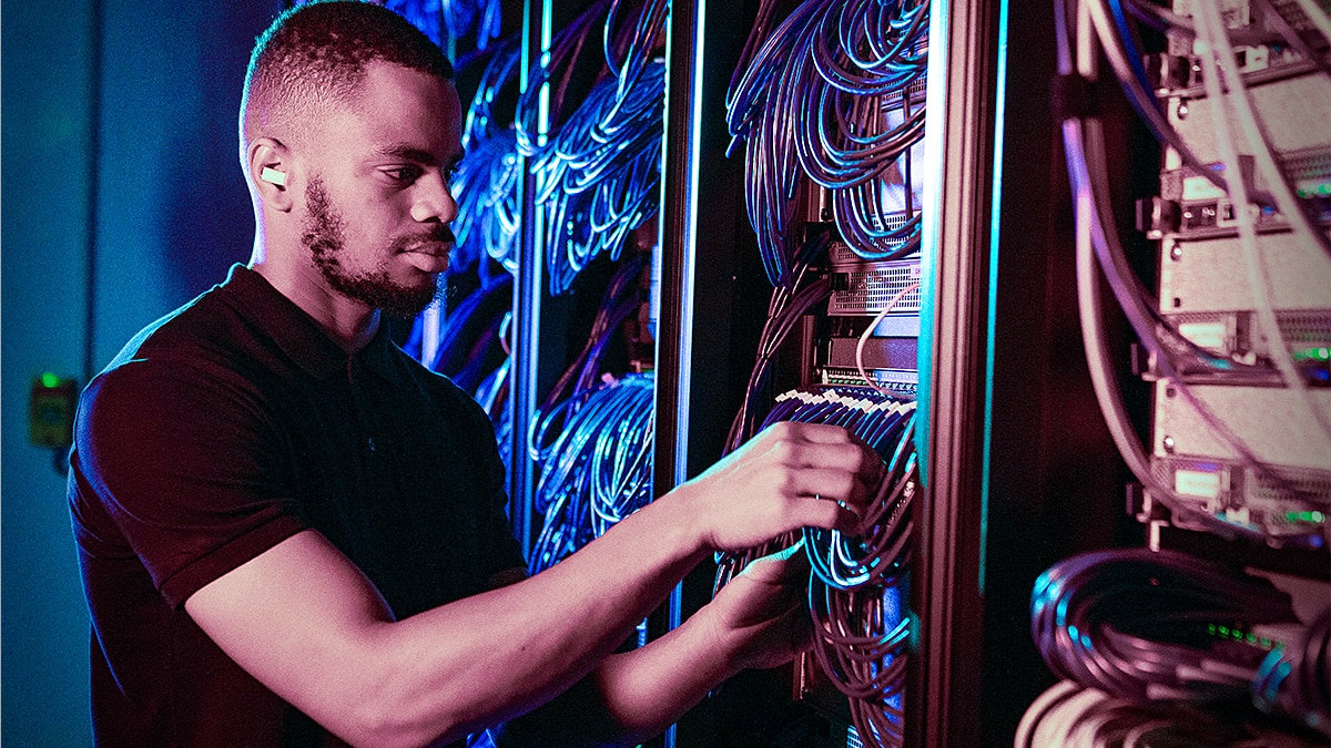 Network technician in a black shirt connecting cables to server rack equipment under blue and purple lighting.