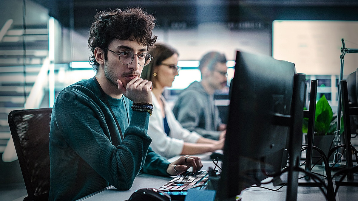 Developer in a teal sweater working at a desktop computer with two colleagues in a modern office.