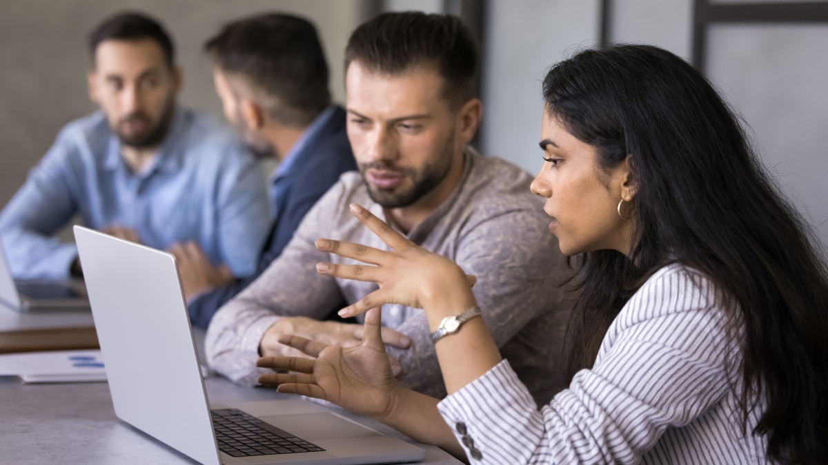 A woman in a striped shirt gestures while discussing with a man over a laptop in an office meeting. Two men are blurred in the background.