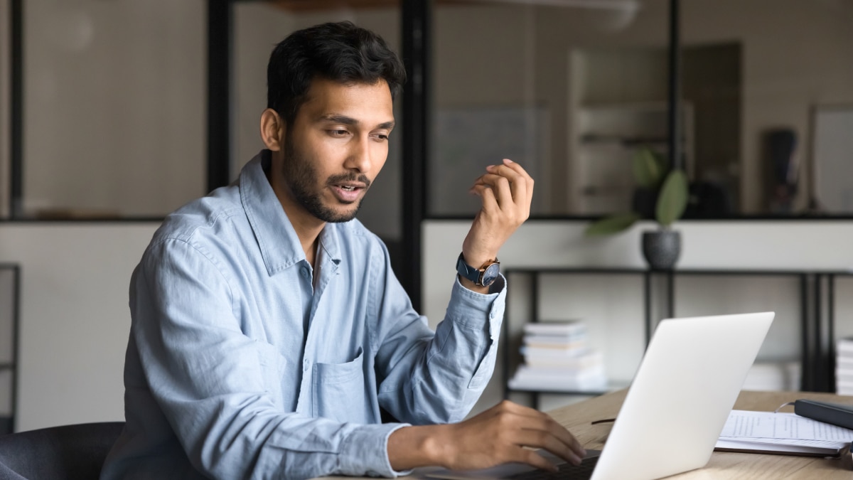Man in blue shirt speaking and gesturing while using a silver laptop at a wooden desk in an office setting.