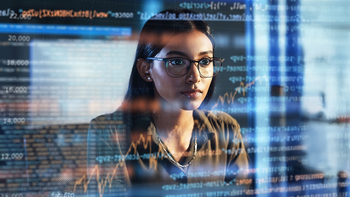 Focused woman in glasses analyzing code and stock market graphs displayed on a transparent screen.