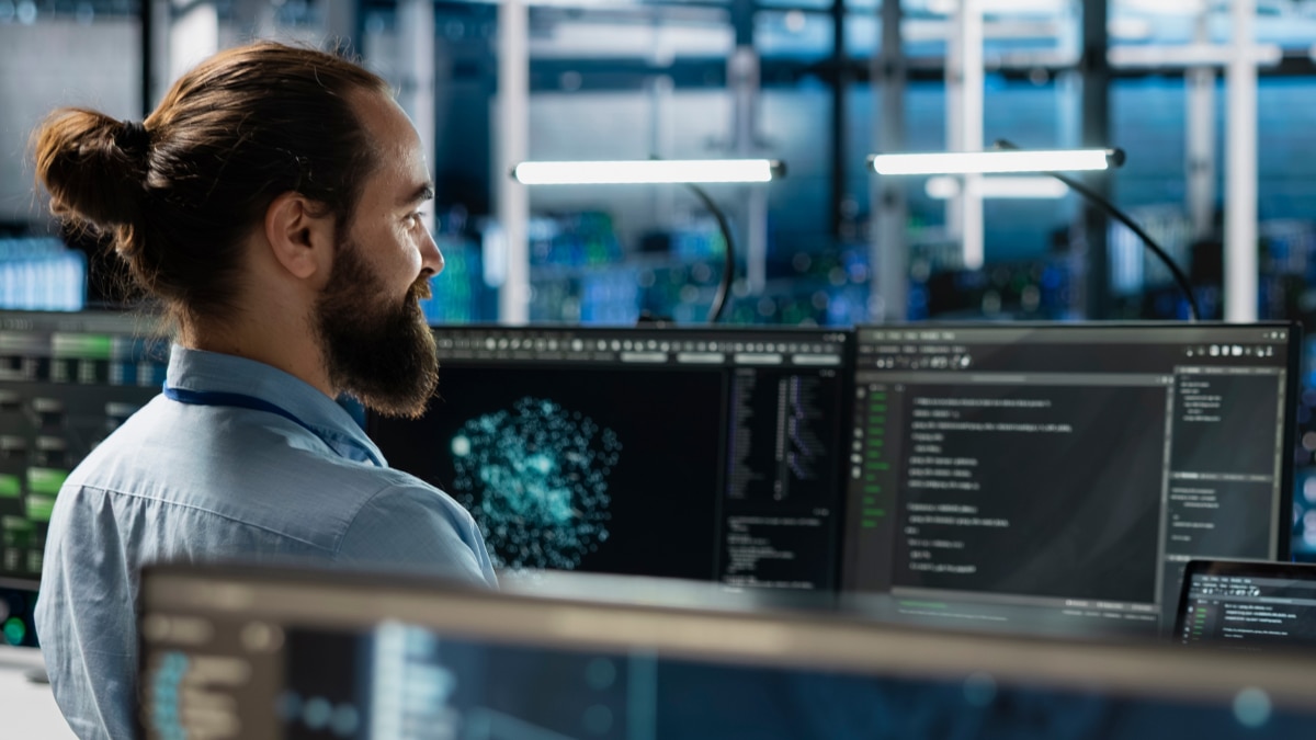 Bearded man in a modern office reviewing data visualizations and programming code on multiple monitors.