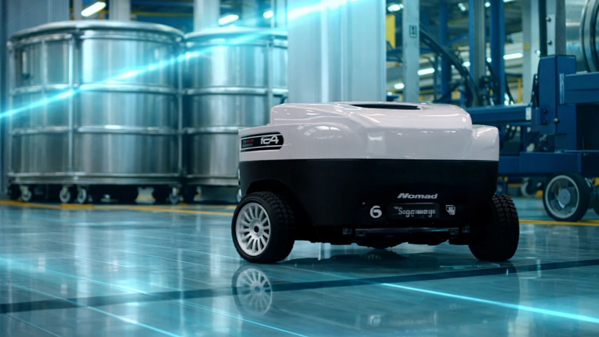 White and black Nomad Segway delivery robot on factory floor with large metal tanks and blue industrial equipment in background