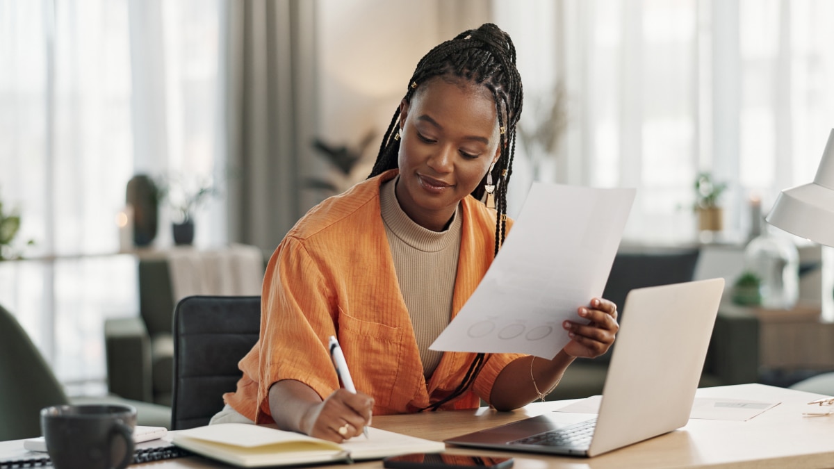 Woman in orange shirt reviews printed document while writing in notebook at desk with laptop and coffee mug