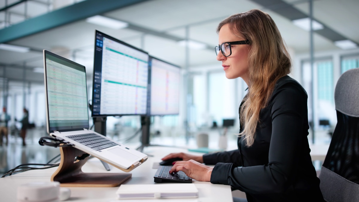 Woman with glasses types at desk with laptop on stand and dual monitors displaying spreadsheets in office