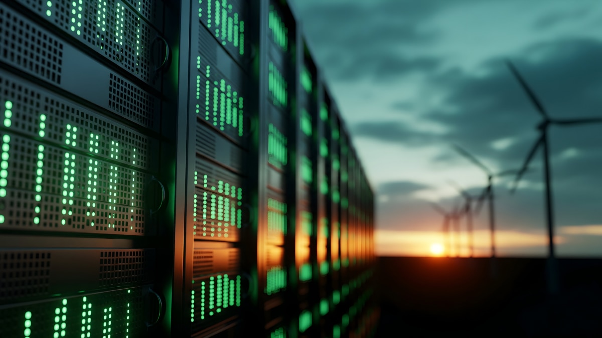 Server racks with green lights beside wind turbines at sunset, representing sustainable data center energy