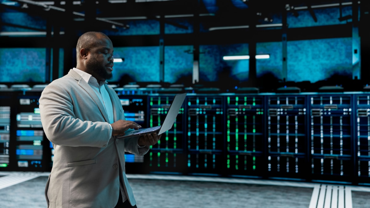 Man using laptop in data center aisle with server racks and blue indicator lights, monitoring systems or infrastructure