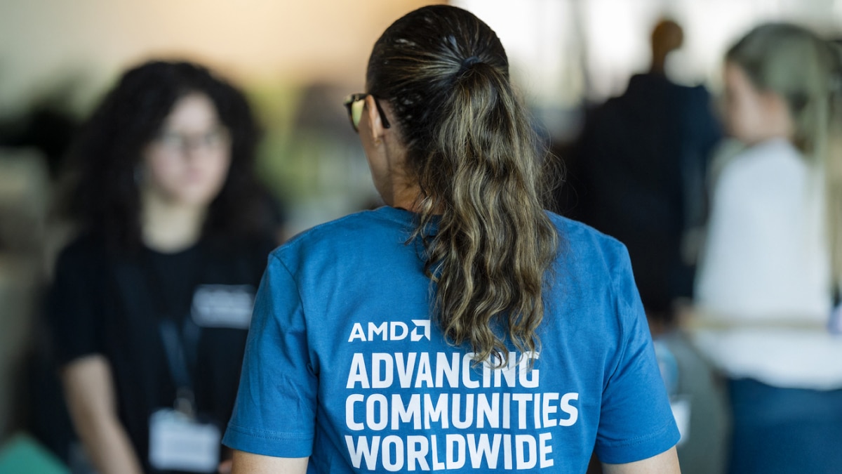Person wearing blue shirt reading “ADVANCING COMMUNITIES WORLDWIDE” with AMD logo, others blurred in indoor event setting.