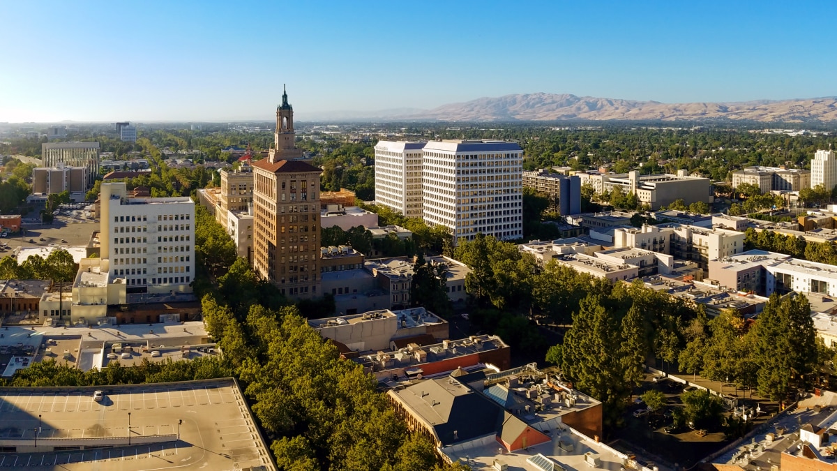 Aerial view of a downtown district with mid-rise office buildings, tree-lined streets, and mountains in the distance under a clear blue sky.