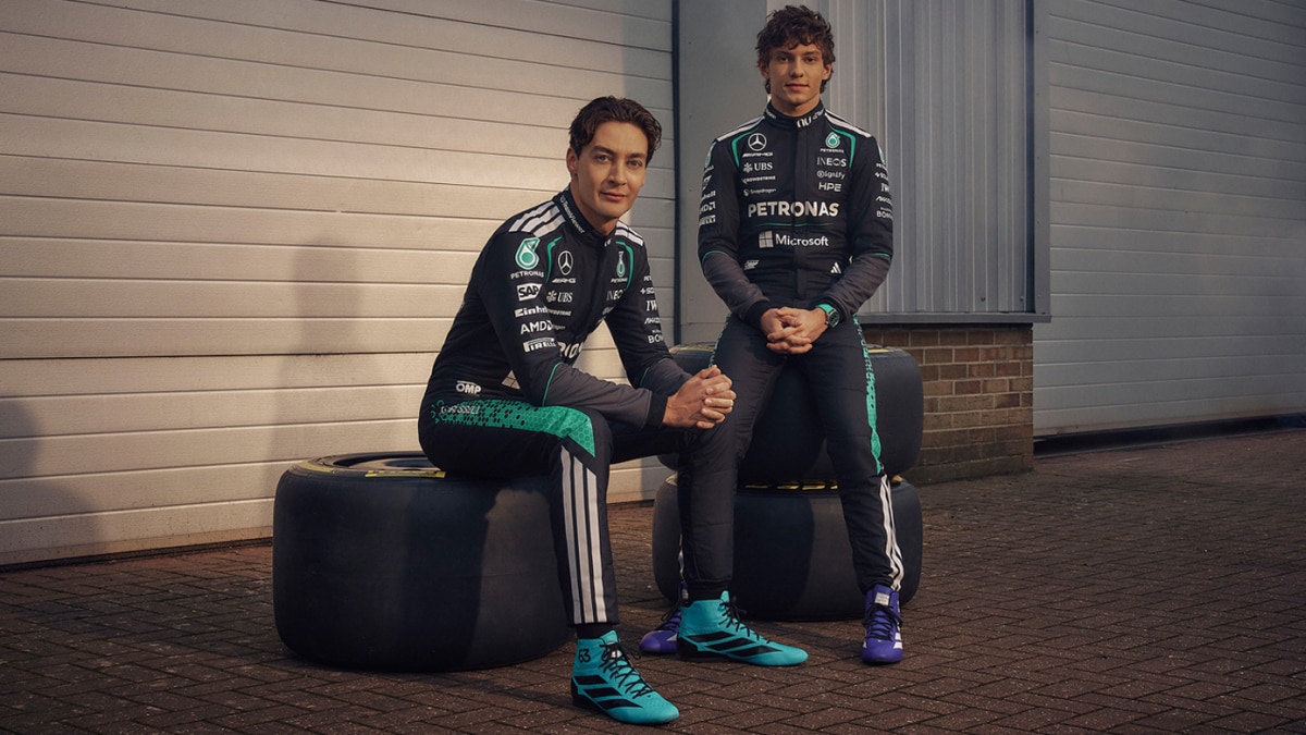 Motorsport driver in black PETRONAS racing suit and teal helmet raises fist on racetrack with sponsor logos visible.