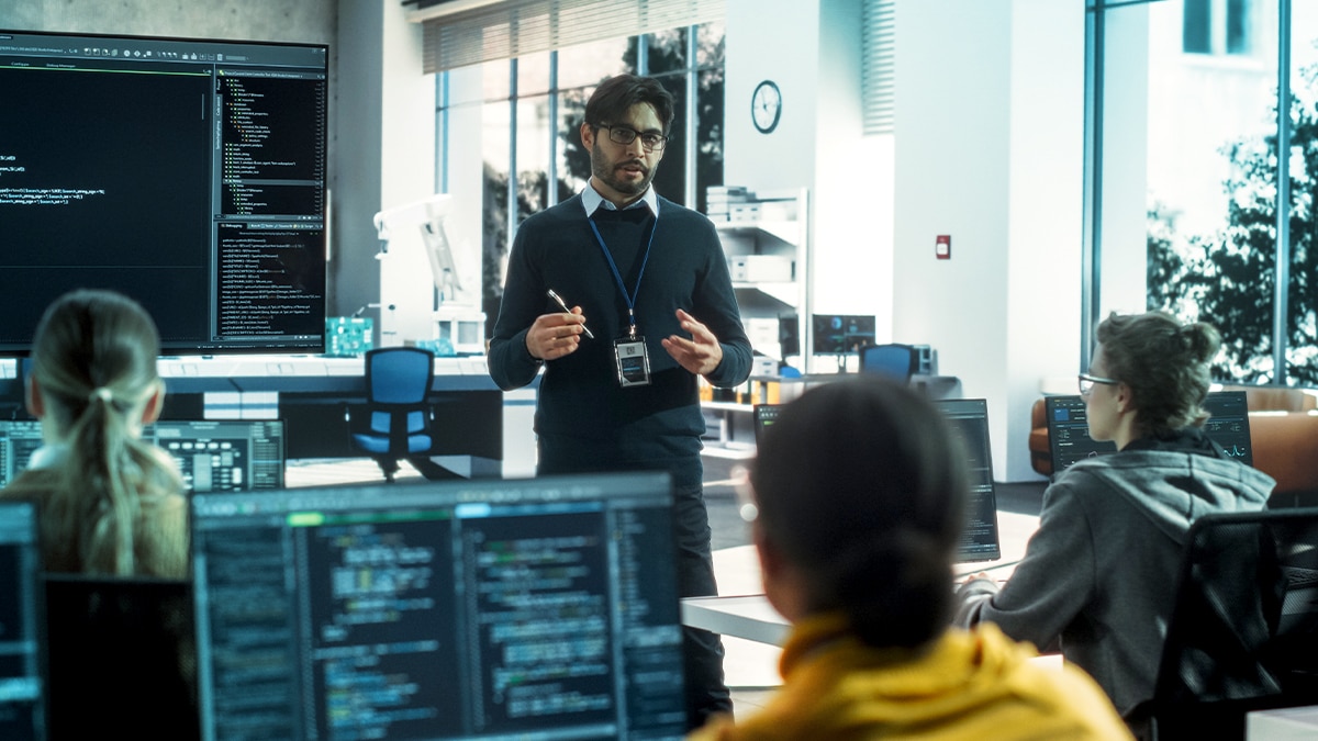 Instructor presenting to students in a classroom, code displayed on monitors during a collaborative programming session.
