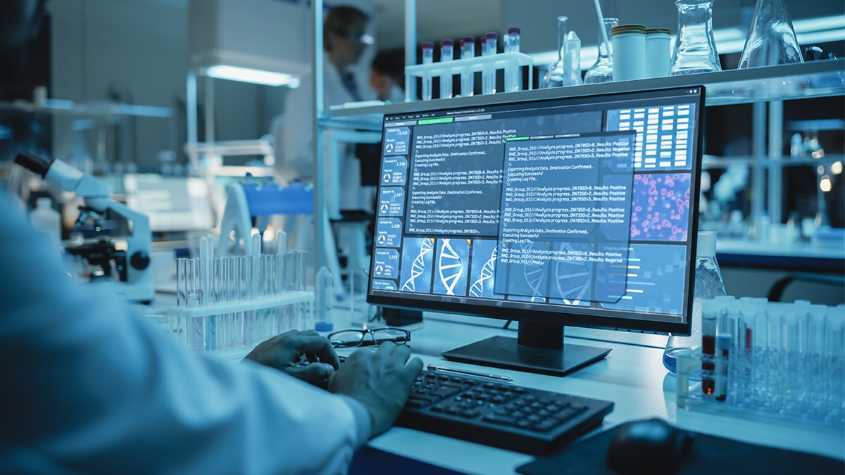 Medical Research Scientist Typing Sophisticated Coding on His Desktop Computer in a Biological Applied Science Research Laboratory. Lab Engineers in White Coats Conduct Experiments in the Background