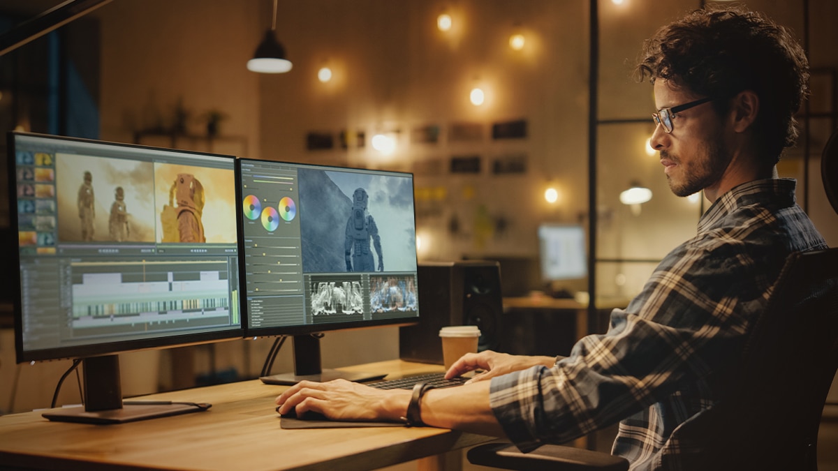 Man editing astronaut footage on dual monitors with color wheels and timeline in warm studio