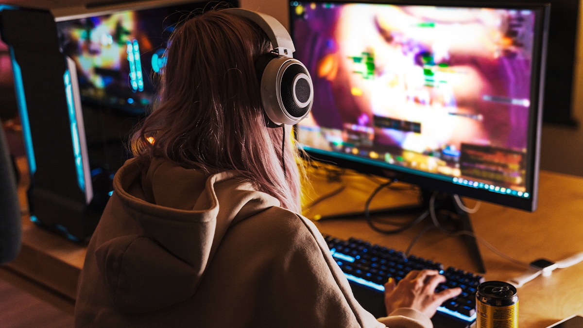 Person wearing over-ear headphones gaming at RGB-lit desktop PC, colorful game on monitor, keyboard and energy drink on desk.