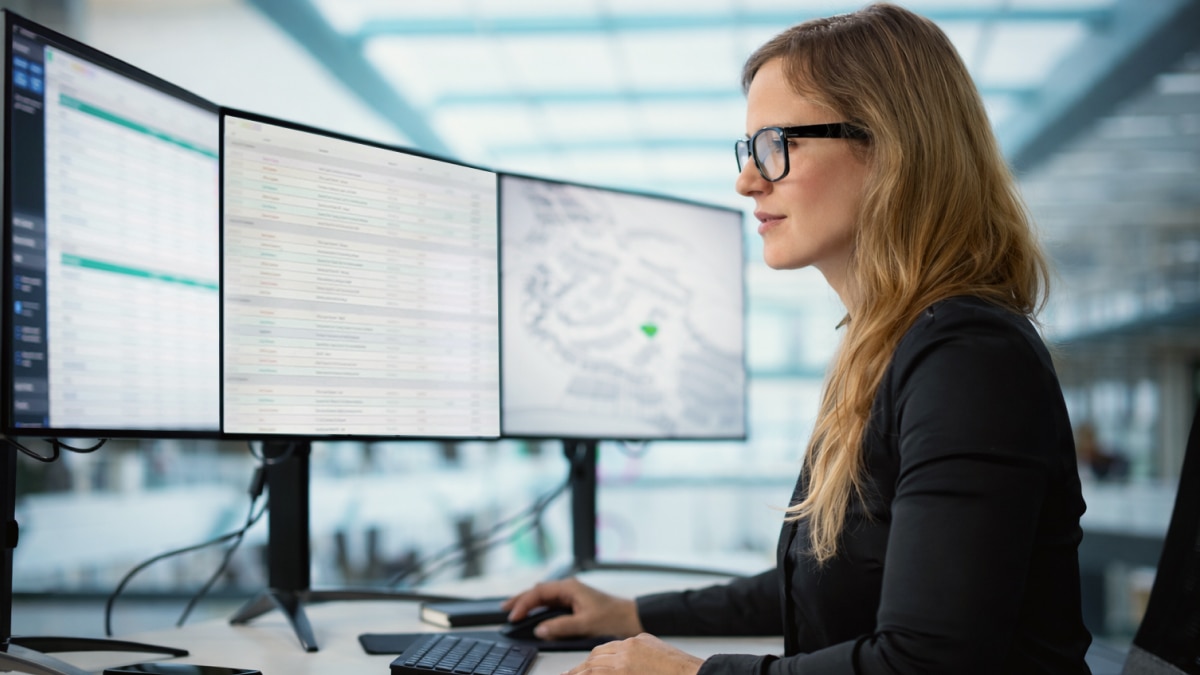 Woman with glasses reviews data dashboard and map on dual monitors in bright modern office