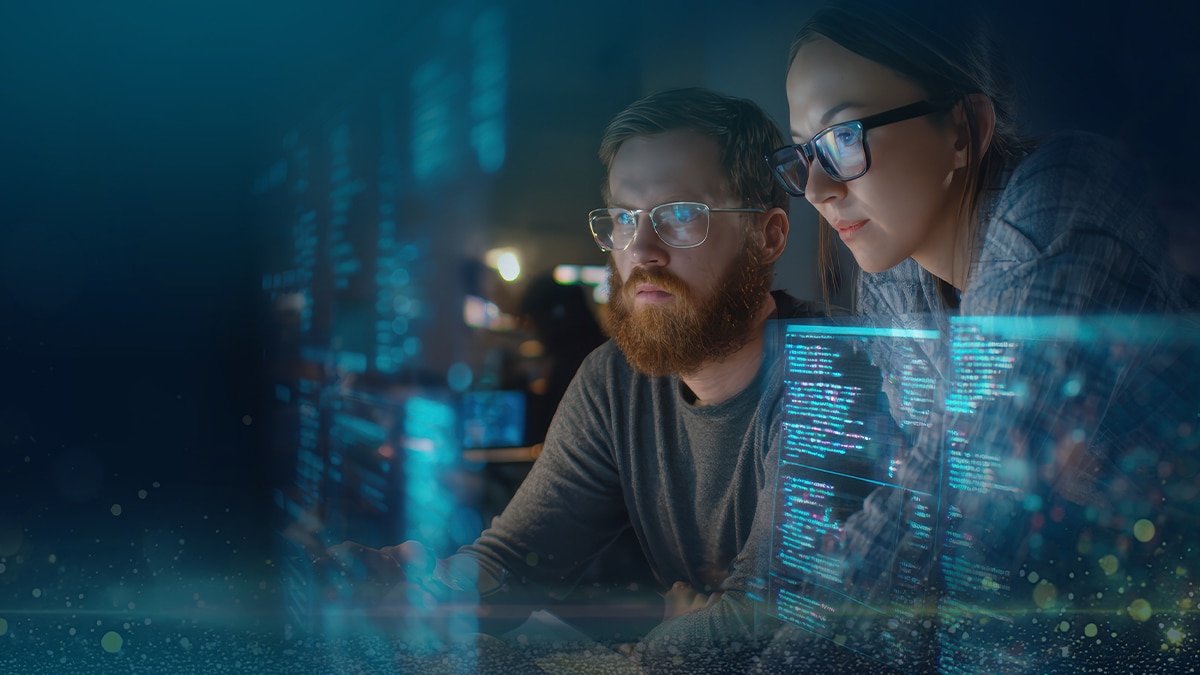 Two developers wearing glasses collaborate at a computer with blue code overlays on screen in a dim workspace