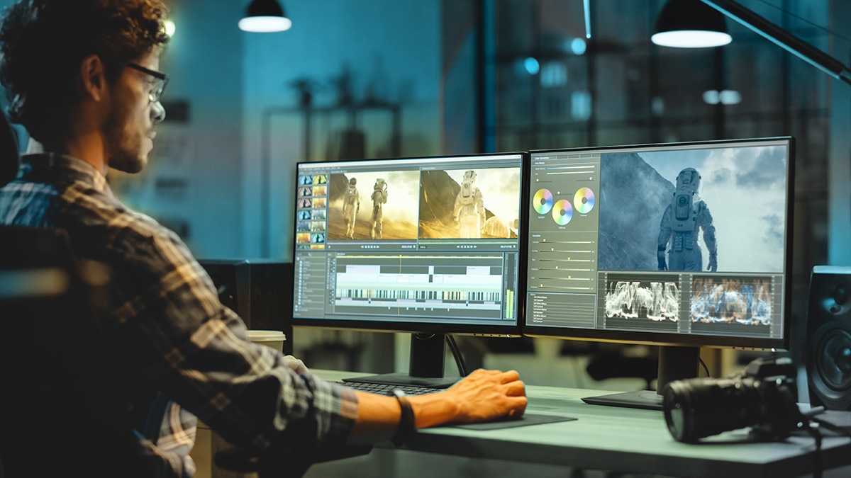 Person editing video at dual monitors showing timeline, color wheels, and astronaut footage in a warmly lit workspace with camera on desk