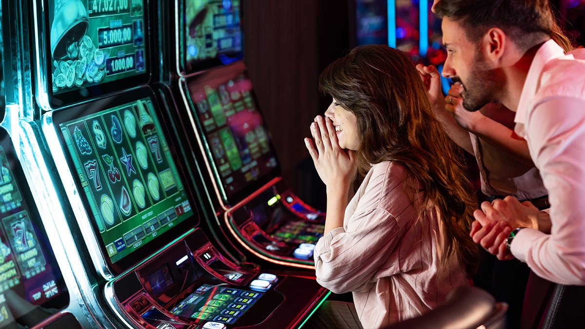 Excited woman and man at slot machines in a casino. Woman smiles with hands to face, man holds her hand, looking at screen showing R$ 47.027,00.
