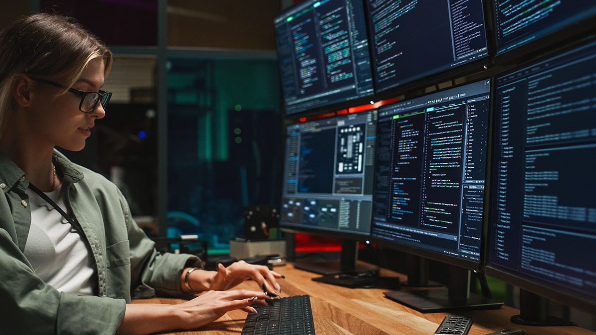 Young woman in glasses and green jacket types on keyboard, looking at multiple monitors displaying lines of code in a dark room.