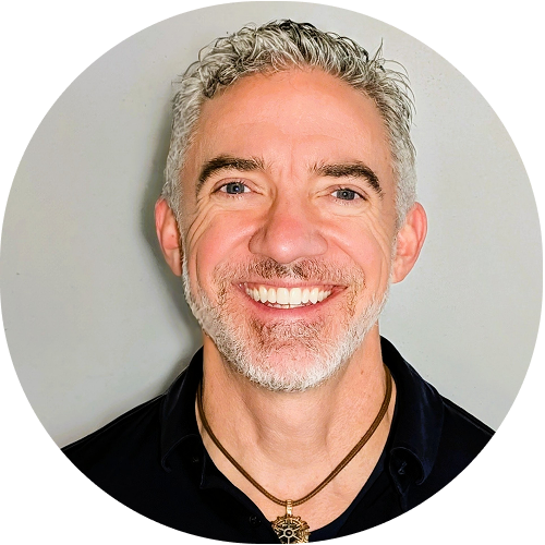 Smiling man with short gray hair and beard wearing a black shirt and pendant against a light gray background.
